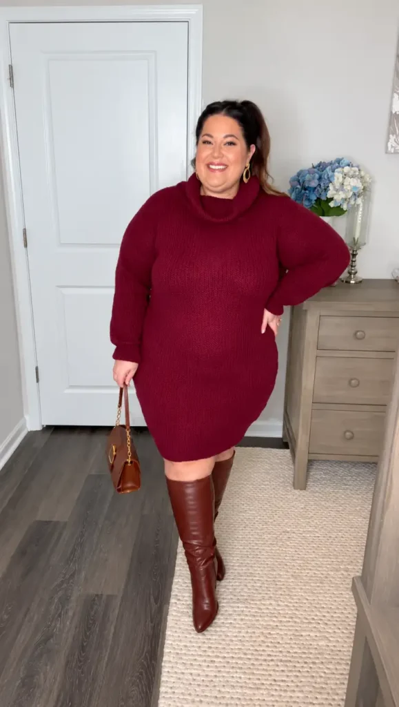 Plus size woman wearing a burgundy turtleneck sweater dress with cognac brown knee-high leather boots, a structured brown handbag, and gold earrings.