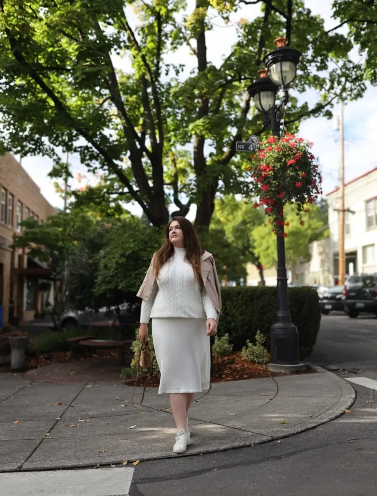 Plus size woman wearing a cream cable-knit sweater with a matching midi skirt, neutral jacket, white sneakers, and a tan handbag for fall.