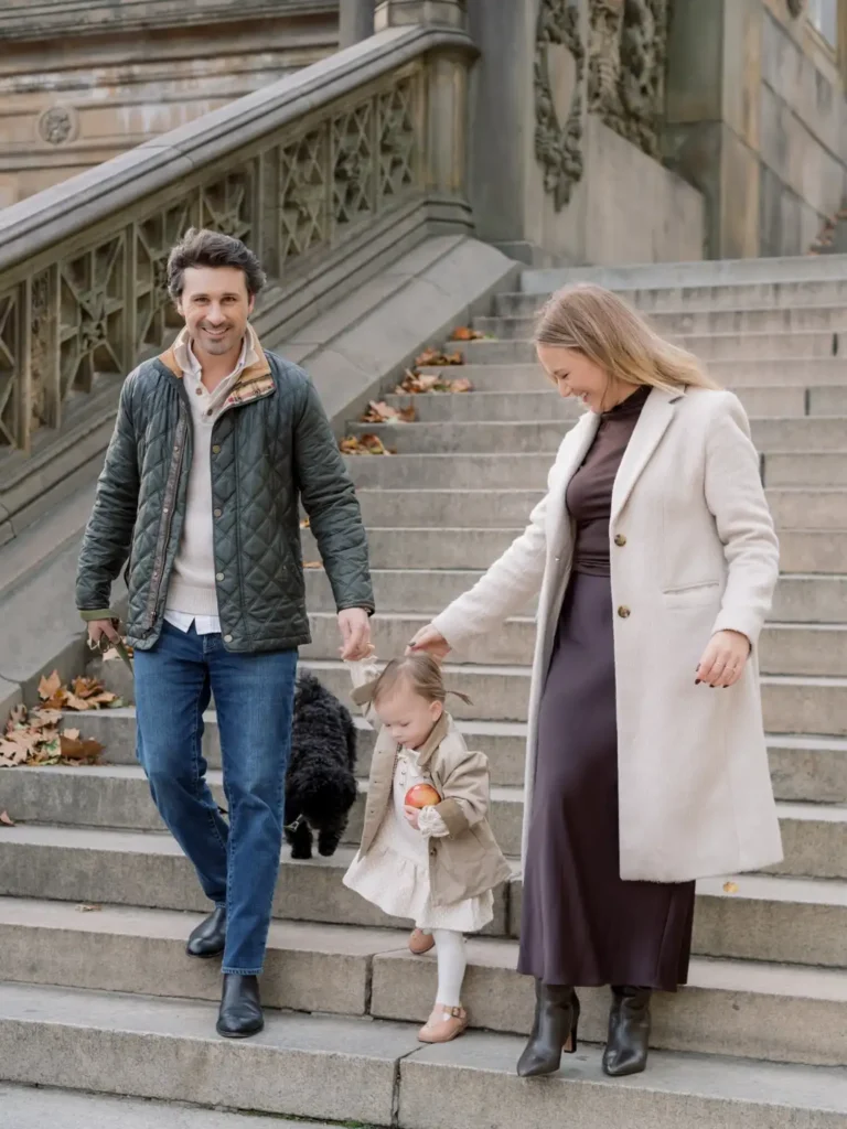 Family wearing layered fall outfits with Mom in a plum dress and ivory coat, Dad in a green quilted jacket and jeans, and their daughter in a beige coat over a cream dress.