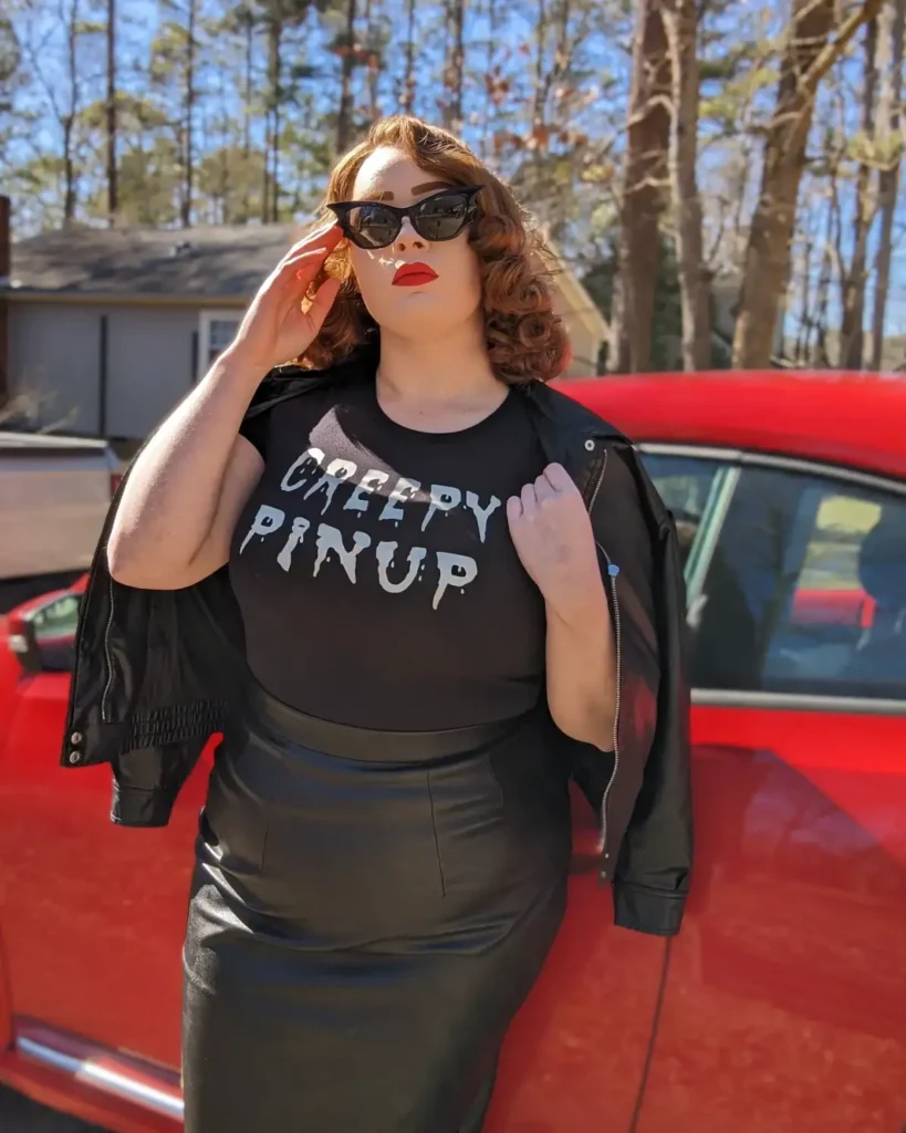 Plus size woman wearing a retro Halloween pinup outfit with a black graphic tee, faux leather skirt, sunglasses, and red lipstick.