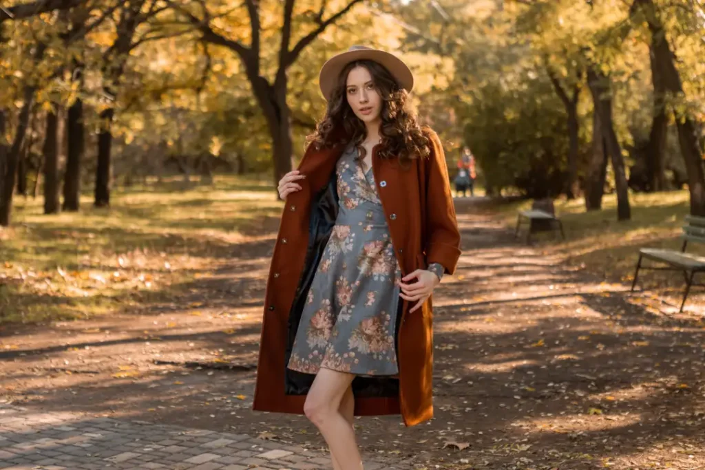 Woman wearing a rust brown long coat over a blue floral mini dress with a beige wide-brim hat for a feminine autumn outfit.