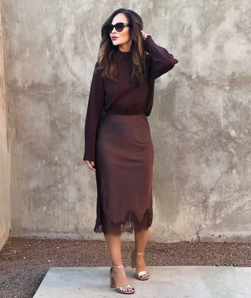 Woman wearing a dark brown sweater with a matching satin midi skirt featuring black lace trim and metallic ankle-strap heels.