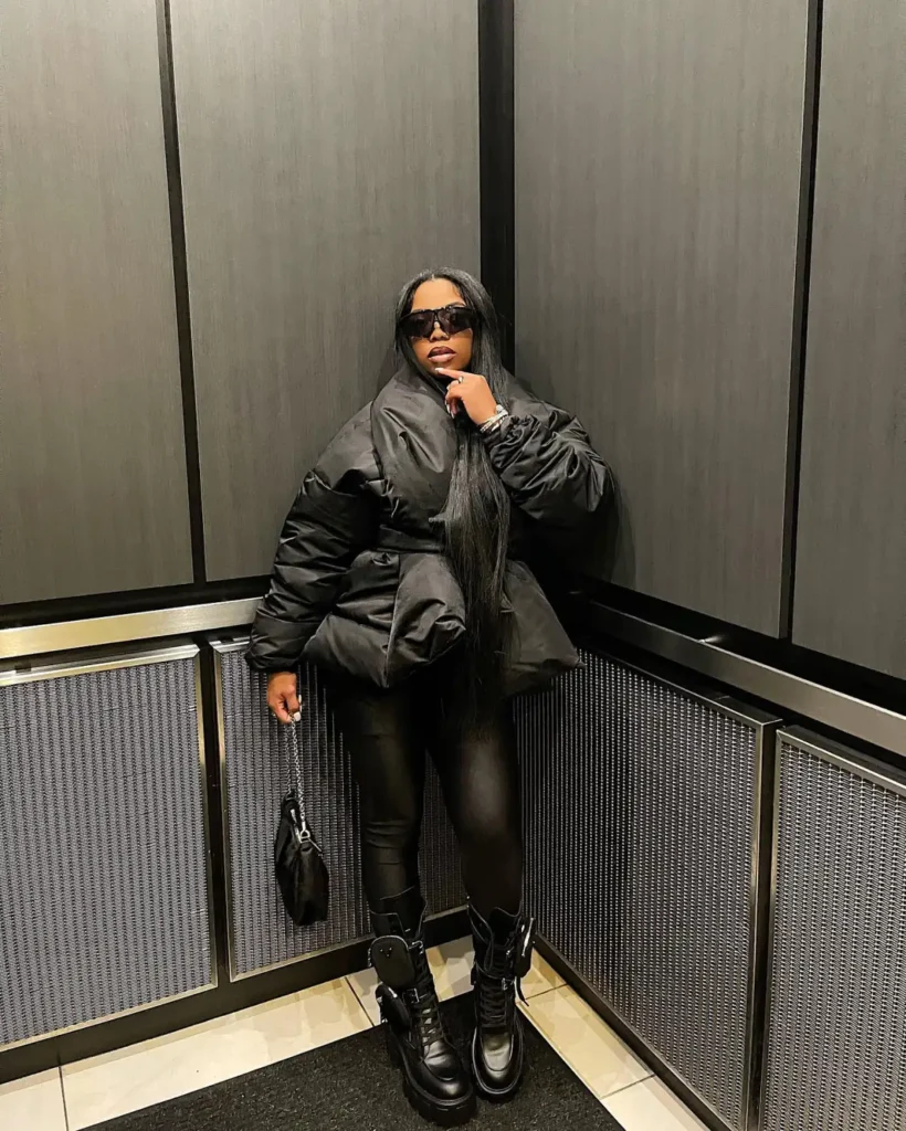 Black woman wearing an oversized black puffer jacket with fitted black pants, chunky lace-up boots, dark sunglasses, and a small black handbag for an edgy fall street-style look.