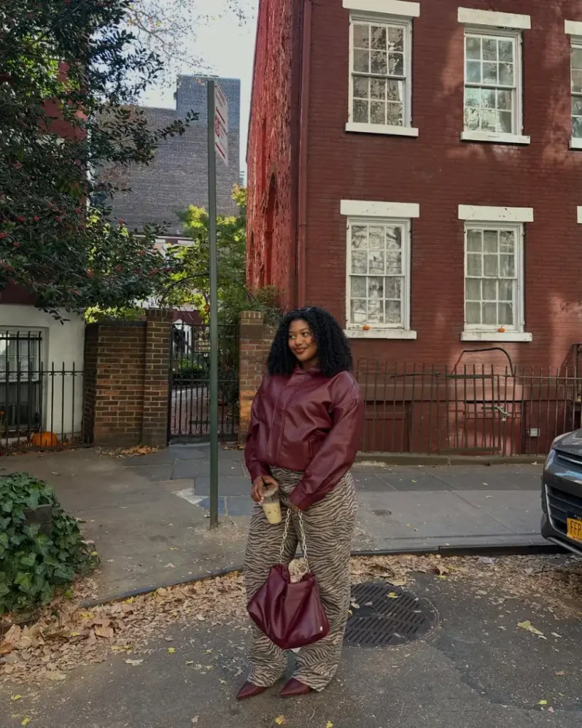Black woman wearing a burgundy leather-look bomber jacket with loose zebra-print trousers, pointed burgundy shoes, and a matching shoulder bag.