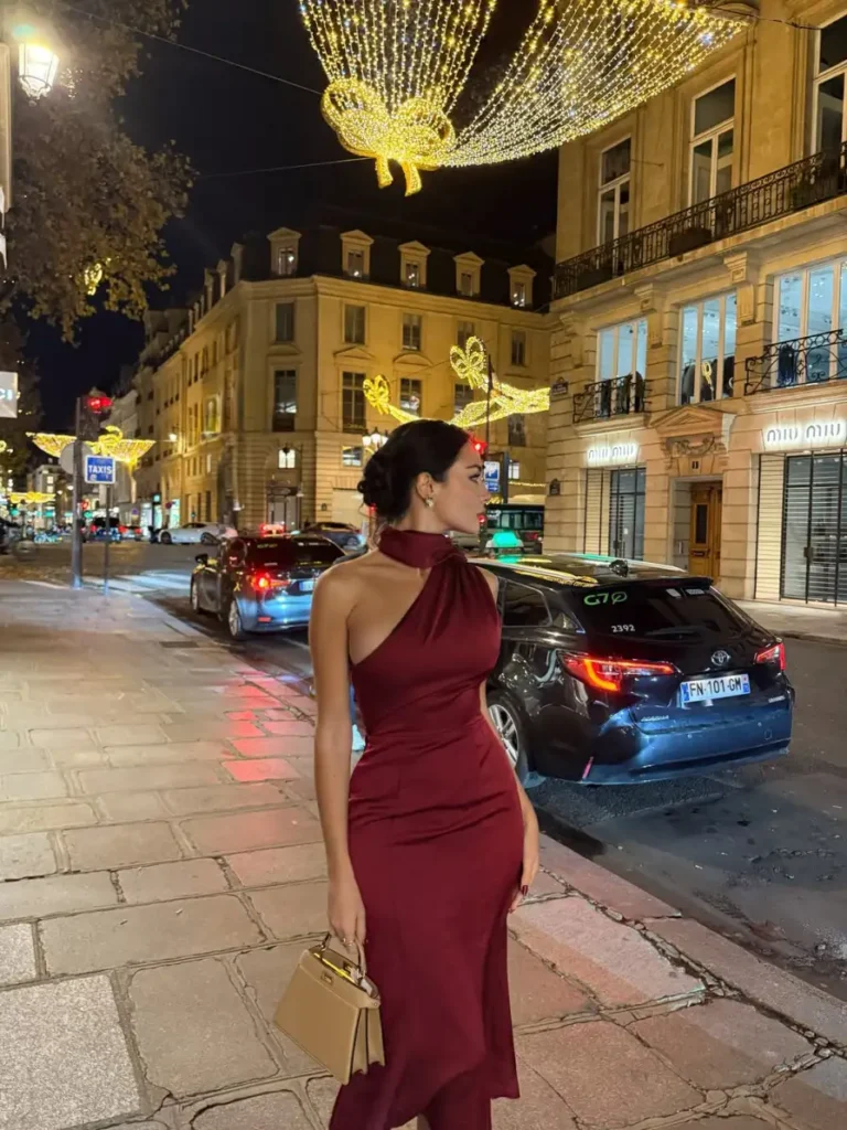 Woman wearing a fitted burgundy asymmetric evening dress with a scarf-style neckline and a small tan top-handle handbag for an elegant fall night look.