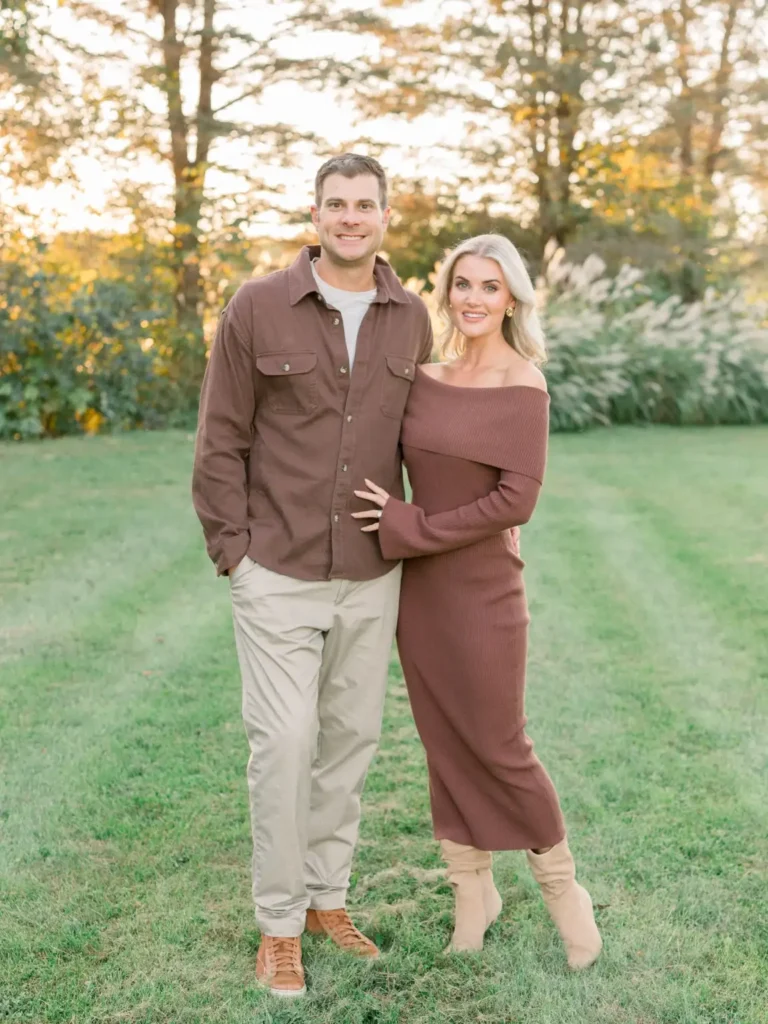 Couple wearing matching chocolate brown fall outfits, with the woman in an off-shoulder ribbed sweater maxi dress and beige boots and the man in a brown overshirt and cream trousers.