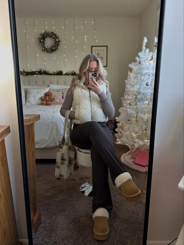 Woman wearing a cream cable-knit zip-up vest over a striped long-sleeve top with dark wide-leg trousers, tan shearling-lined slippers, and a patterned tote for a casual cozy outfit.