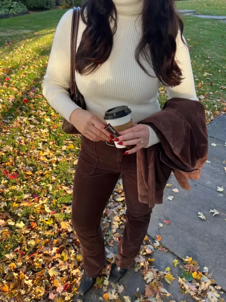 Woman wearing a cream ribbed turtleneck with chocolate brown trousers, pointed black shoes, and a brown jacket for a classic casual fall outfit.