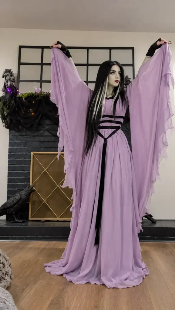 Woman wearing a flowing lavender gothic gown with oversized chiffon sleeves, black waist straps, long dark hair, and dramatic makeup.