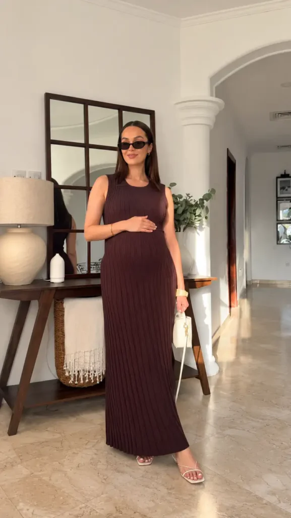 Pregnant woman wearing a sleeveless chocolate brown ribbed maxi dress with white strappy sandals, gold jewelry, and an ivory handbag for an elegant maternity Thanksgiving look.