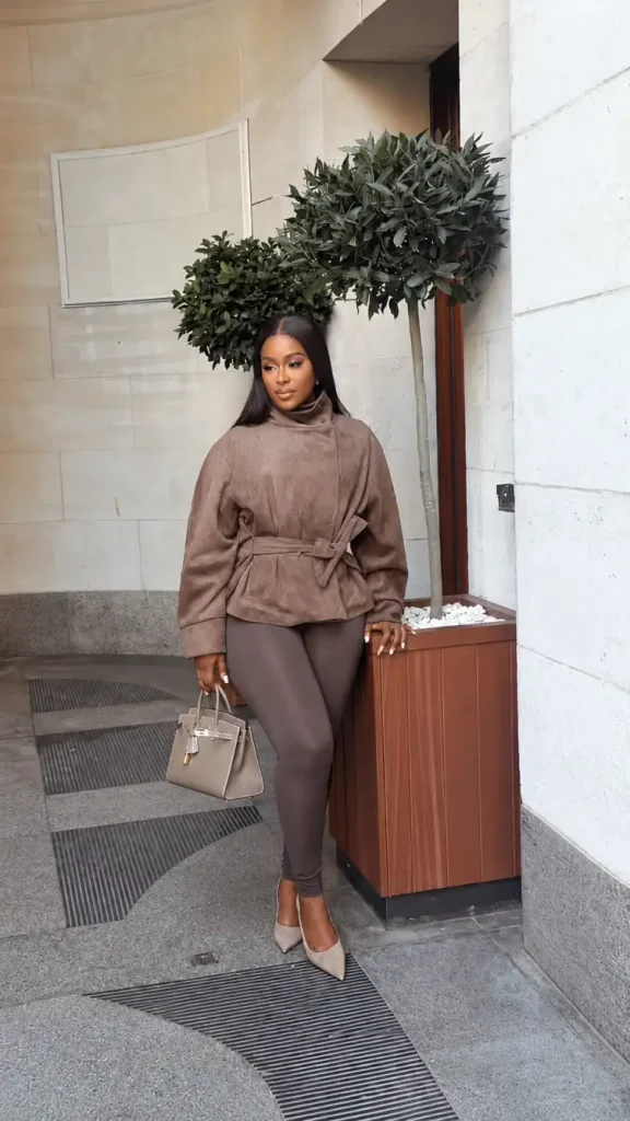 Black woman wearing a belted mocha suede-look jacket with fitted brown trousers, nude pointed pumps, and a structured taupe handbag for sophisticated autumn office style.
