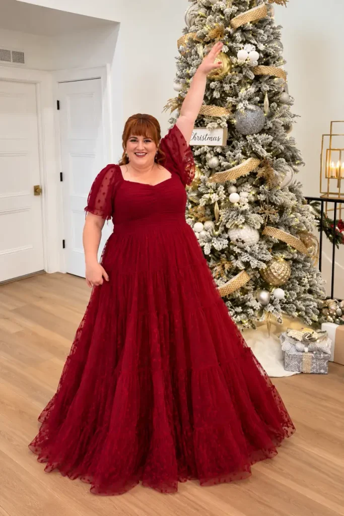 Plus-size woman wearing a burgundy red floor-length tulle ballgown with a ruched bodice, sheer puff sleeves, and a tiered skirt for a formal Christmas celebration.