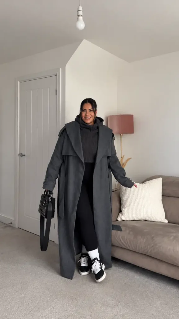 Plus size woman wearing a long charcoal gray coat over a chunky turtleneck sweater with black leggings, white ribbed socks, and black-and-white sneakers.