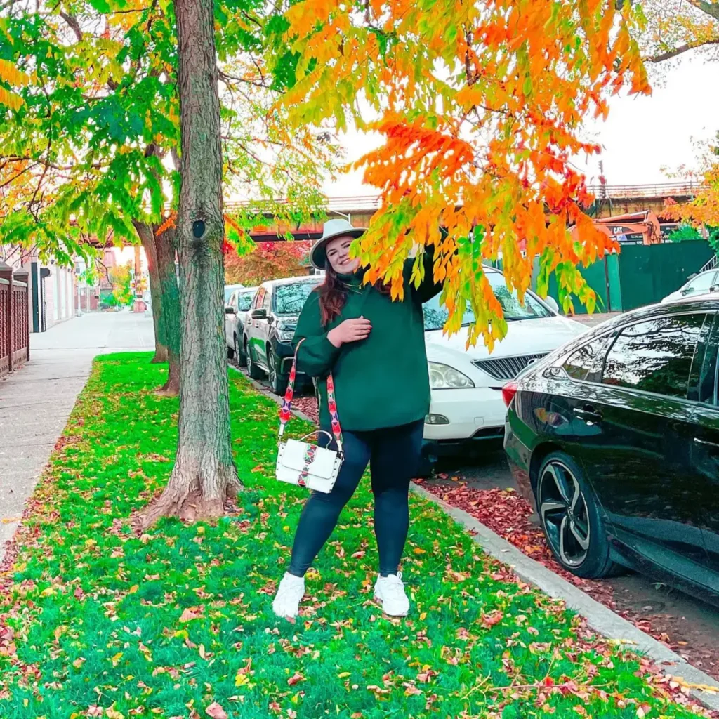 Plus size woman wearing an oversized forest green sweater with black leggings, white sneakers, a wide-brim hat, and a structured white handbag for fall.