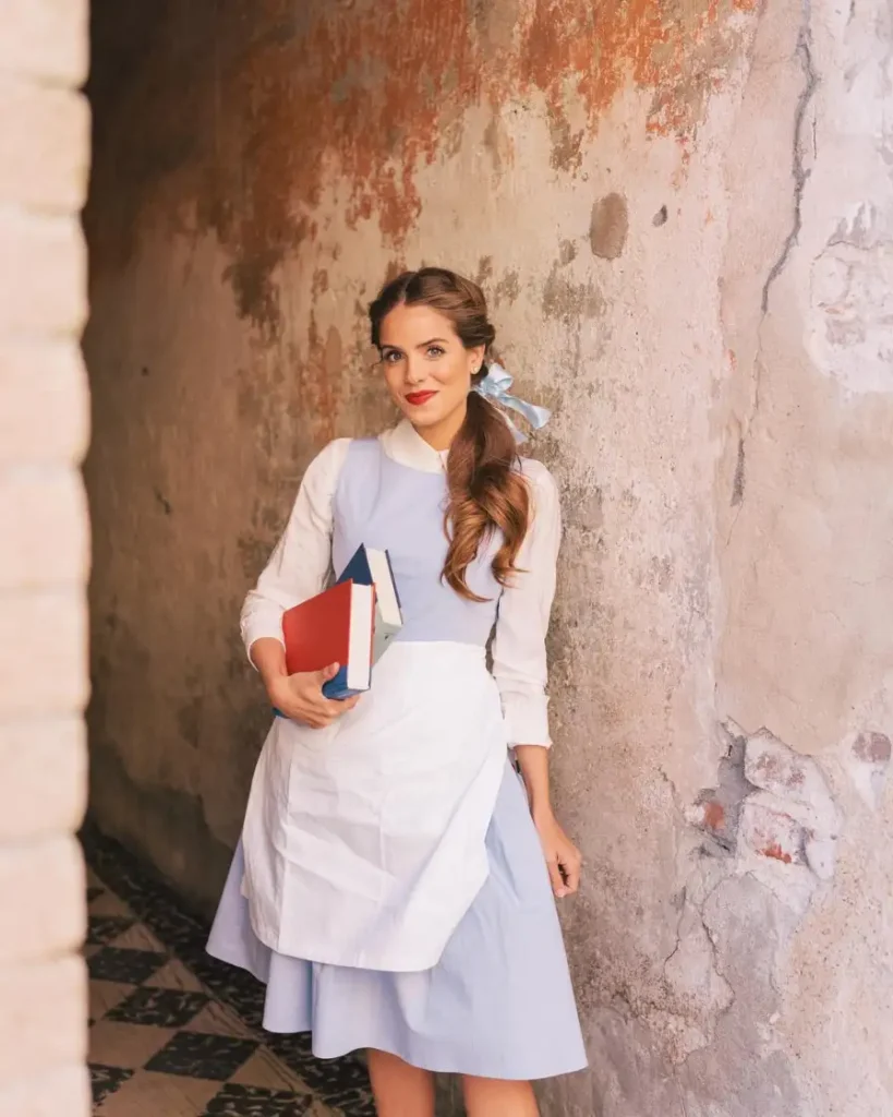 Woman wearing a pale blue storybook dress with white blouse and apron, blue hair ribbon, and books for a classic Halloween costume.