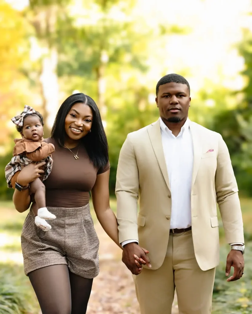 Family wearing coordinated beige and brown outfits with Dad in a cream suit, Mom in a fitted brown top and checked shorts, and their baby in a brown outfit with plaid details.