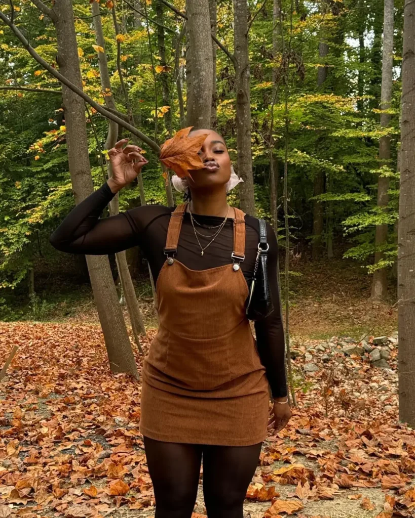 Black woman wearing a brown corduroy pinafore mini dress over a sheer black long-sleeve top with black tights and a small shoulder bag for an earthy fall outfit.