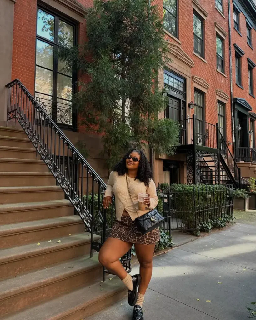 Black woman styling a cream button-front cardigan with a leopard-print mini skirt, black chunky loafers, beige socks, and a quilted shoulder bag.