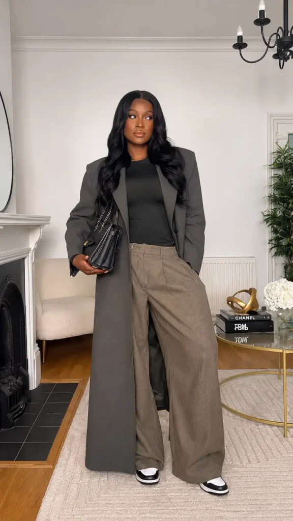 Black woman wearing a long charcoal gray coat over a black top with taupe wide-leg trousers, black-and-white sneakers, and a black handbag for casual fall workwear.