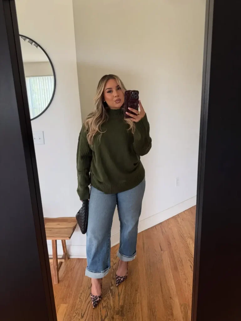 Woman wearing a dark olive green knit sweater with cuffed light-wash jeans, leopard print pointed-toe heels, and a black woven handbag for a chic Thanksgiving outfit.