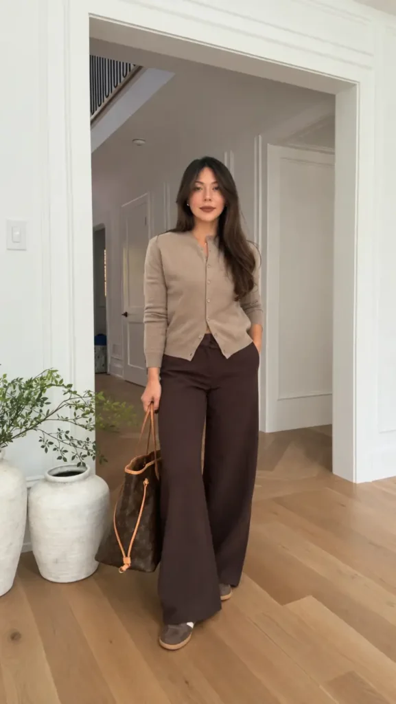 Petite woman wearing a fitted taupe cardigan with chocolate brown wide-leg trousers, neutral sneakers, and a large brown tote bag.