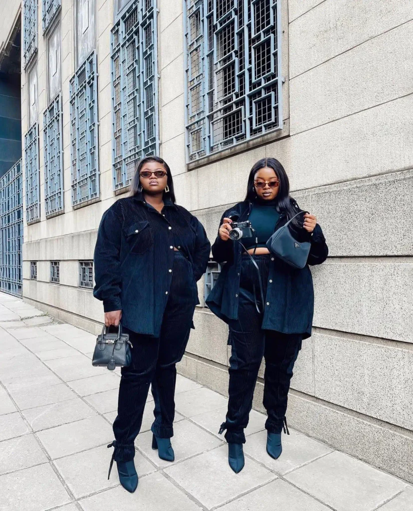 Two plus size Black women wearing coordinated dark navy corduroy shirts and trousers with pointed blue ankle boots, black handbags, and sunglasses for trendy fall street style.