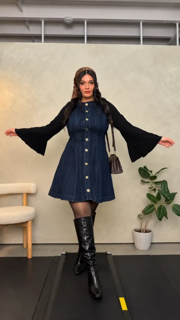 Mid size woman wearing a dark blue button-front denim mini dress over a black flared-sleeve top with sheer tights, knee-high boots, and a brown headscarf.