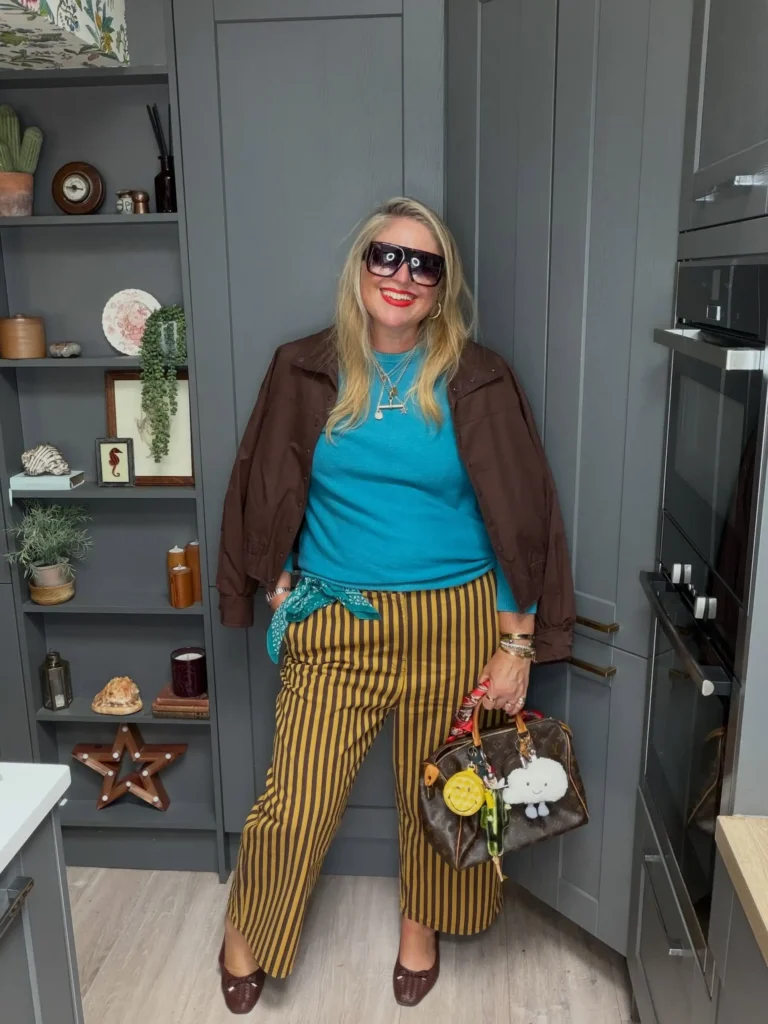 Plus size woman wearing a teal sweater with mustard and brown striped trousers, a cropped brown jacket, brown flats, and a statement handbag for fall.