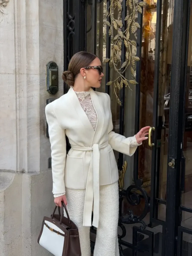 Woman wearing a cream belted blazer over a lace top with textured cream trousers and a structured brown and white handbag.