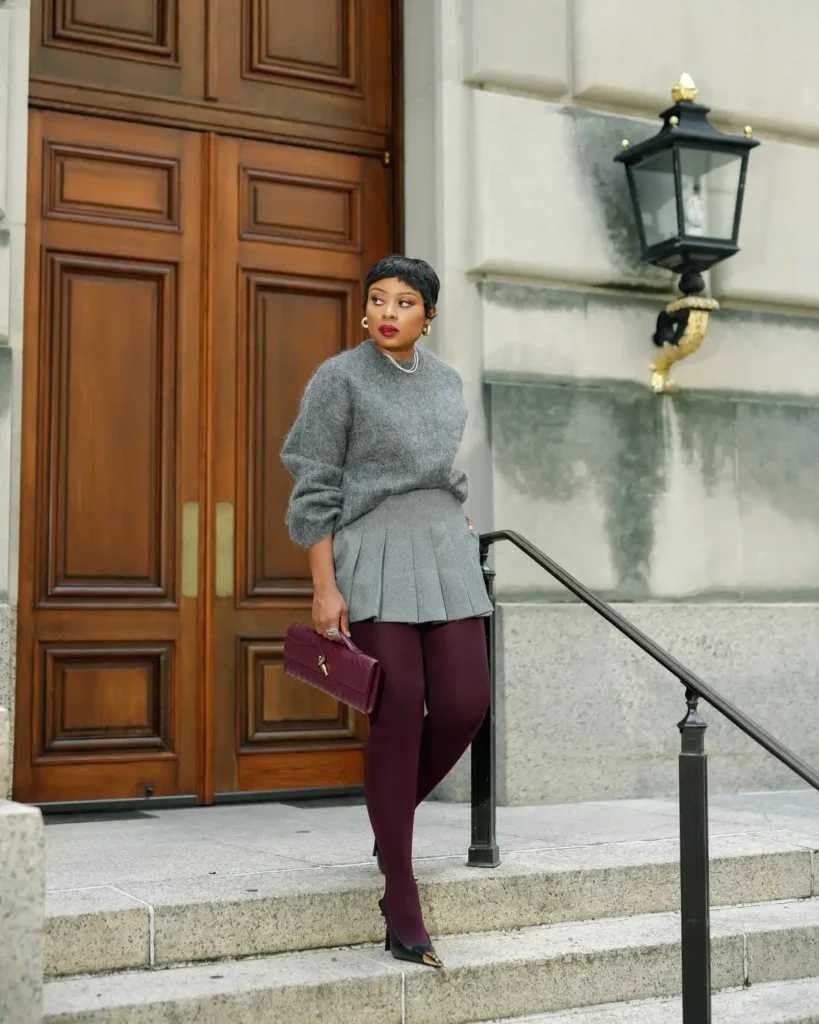 Black woman wearing a soft gray sweater with a matching pleated mini skirt, burgundy tights, pointed black heels, and a burgundy clutch for a polished fall look.