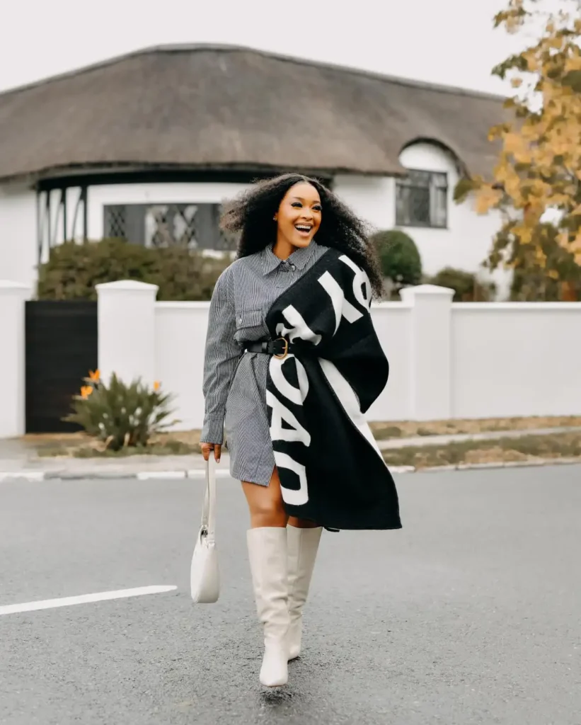 Black woman wearing a black-and-white striped shirt dress with an oversized black graphic scarf, a black waist belt, white knee-high boots, and a white shoulder bag.
