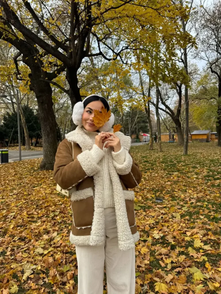 Woman wearing a brown shearling jacket over a cream cable-knit sweater with light trousers and white earmuffs for a cozy cold-weather fall look.