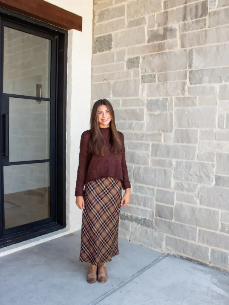 Woman wearing a burgundy chunky knit sweater with a brown plaid midi skirt and brown ankle boots for a cozy, casual autumn Friendsgiving outfit.