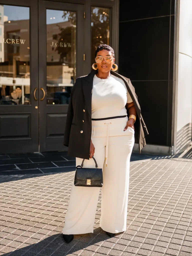 Black woman wearing a cream top with matching wide-leg trousers, a black blazer draped over her shoulders, black pointed heels, and gold hoop earrings.