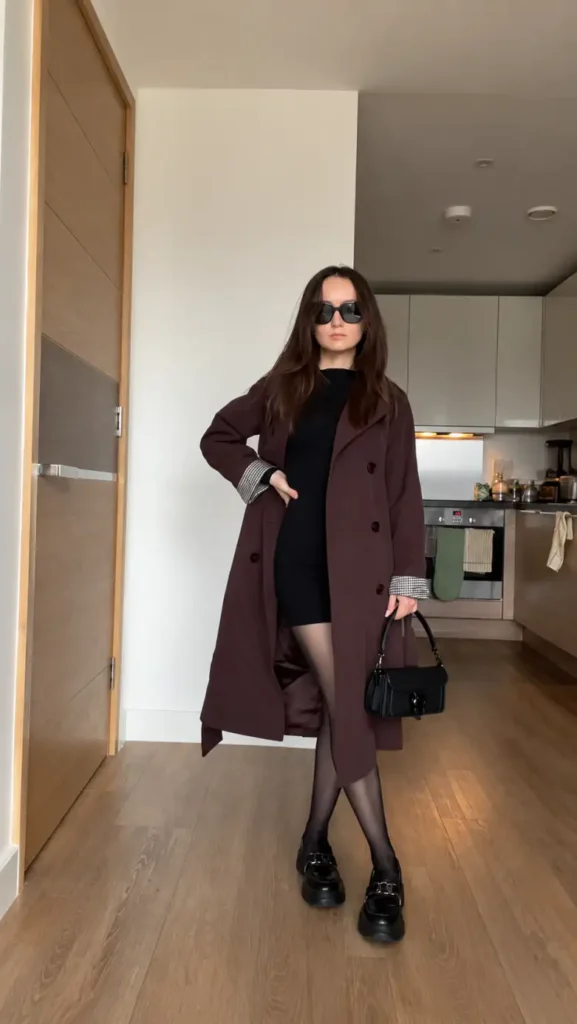 Petite woman wearing a long burgundy coat over a fitted black mini dress with sheer tights, chunky black loafers, and a small black handbag.