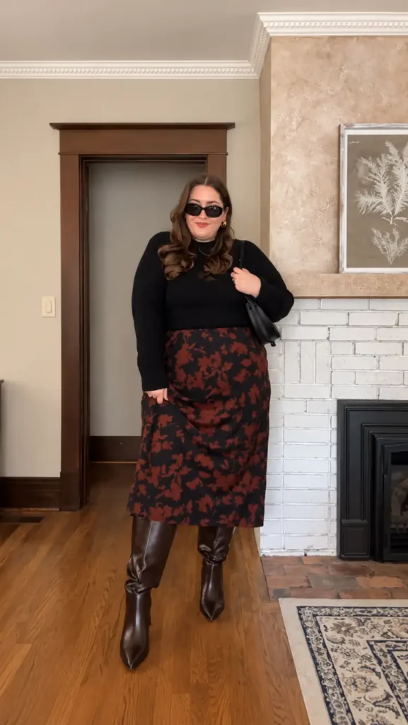 Plus size woman wearing a black fitted sweater with a rust floral midi skirt, brown knee-high boots, sunglasses, and a black shoulder bag for fall.