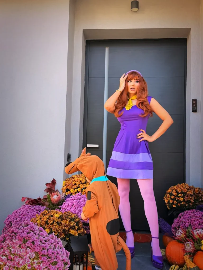Woman wearing a purple mini dress with lavender trim, yellow neck scarf, pink tights, purple heels, headband, and long red hair.