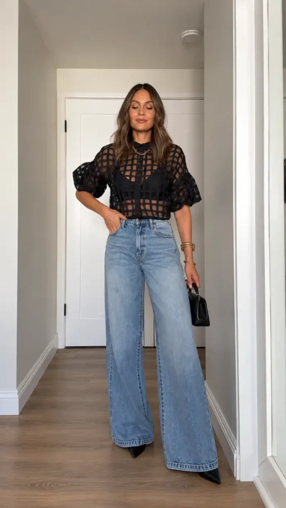 Woman wearing a sheer black grid-pattern blouse with puff sleeves, light blue wide-leg jeans, black pointed heels, and a mini handbag for a dressy casual Thanksgiving outfit.
