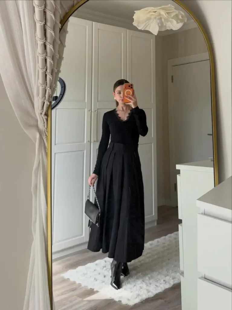 Woman wearing a fitted black lace-trim top with a long pleated black skirt, pointed boots, and a small shoulder bag for an elegant monochrome fall outfit.