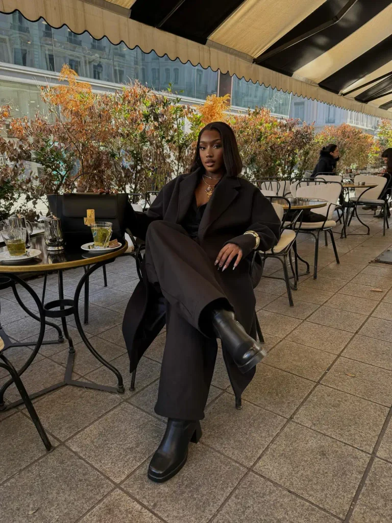Black woman wearing an oversized black tailored coat with matching wide-leg trousers, chunky black ankle boots, and a structured handbag for a minimalist autumn outfit.