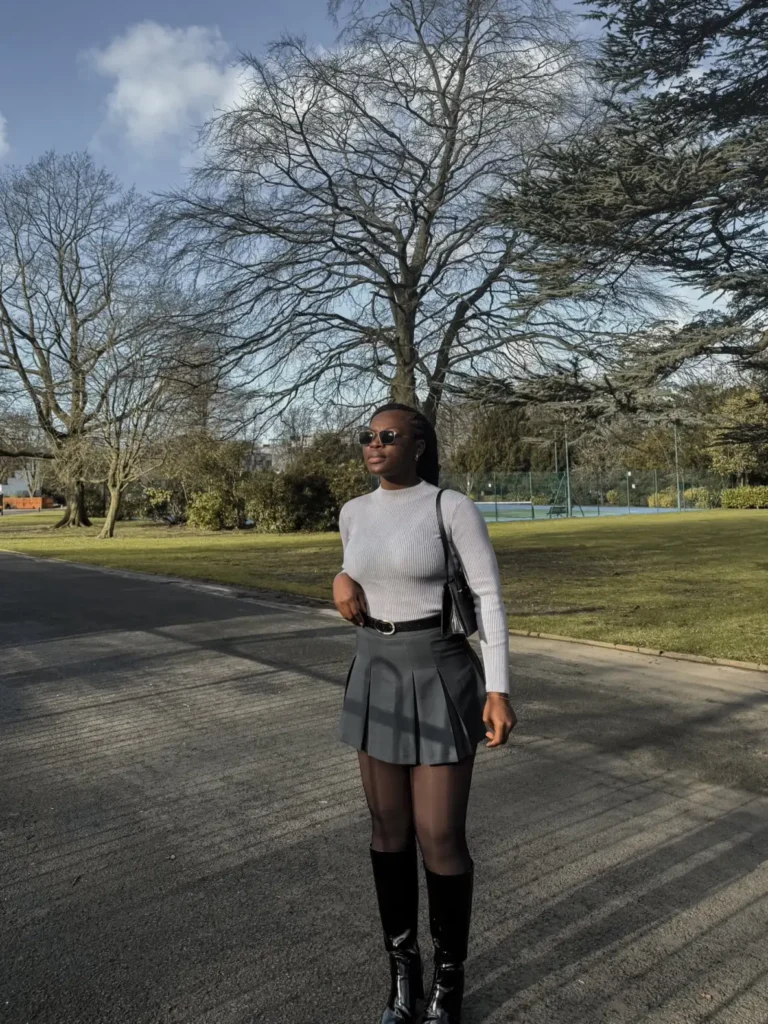 Black woman wearing a fitted light gray ribbed top with a dark pleated mini skirt, sheer tights, glossy black knee-high boots, sunglasses, and a black shoulder bag.