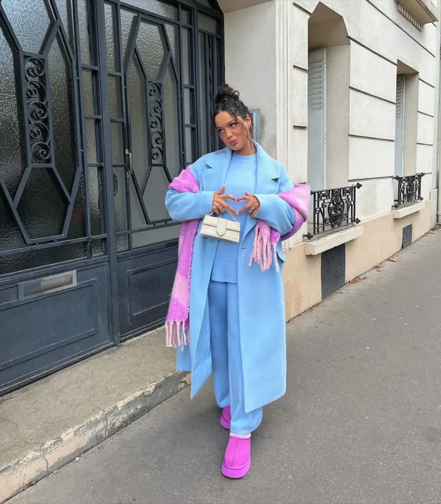 Black woman wearing a pastel blue long coat over a matching blue sweater and wide-leg trousers, accessorized with a bright pink fringed scarf, pink platform boots, and a cream bag.