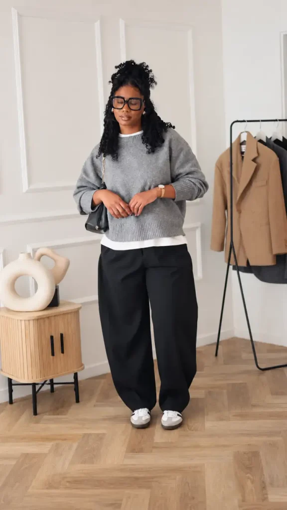 Black woman wearing a relaxed gray sweater over a white shirt with black wide-leg trousers, gray-and-white sneakers, and a black shoulder bag for smart casual office fashion.