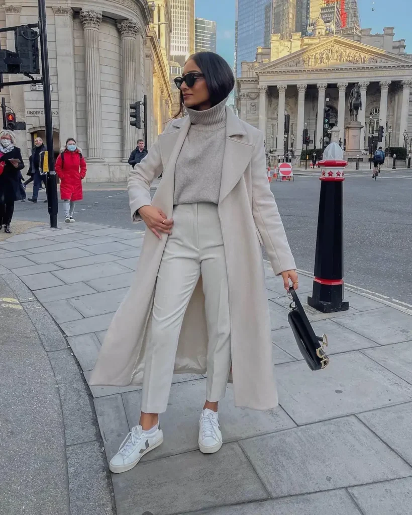 Woman styling a long ivory coat over a gray turtleneck sweater, cream tailored ankle trousers, and white sneakers with a black handbag for a casual chic winter outfit.