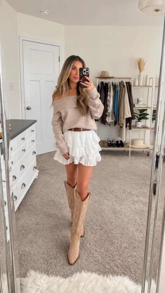 Woman wearing an off-shoulder oatmeal sweater with a white tiered ruffle mini skirt, brown belt, and beige western boots for a cute Friendsgiving outfit.
