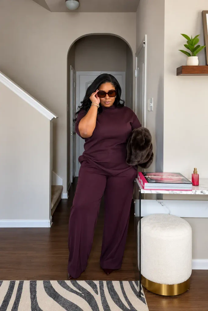 Plus size Black woman wearing a matching deep plum short-sleeve top and wide-leg trousers with burgundy pointed shoes, sunglasses, and a plush brown accessory for a classy fall look.