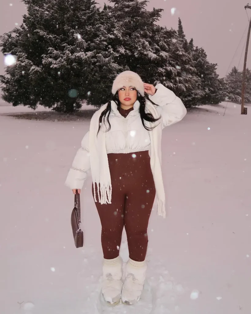 Plus size woman wearing a cropped white puffer jacket with burgundy leggings, a fluffy cream hat, a long white scarf, and warm snow boots outdoors in falling snow.