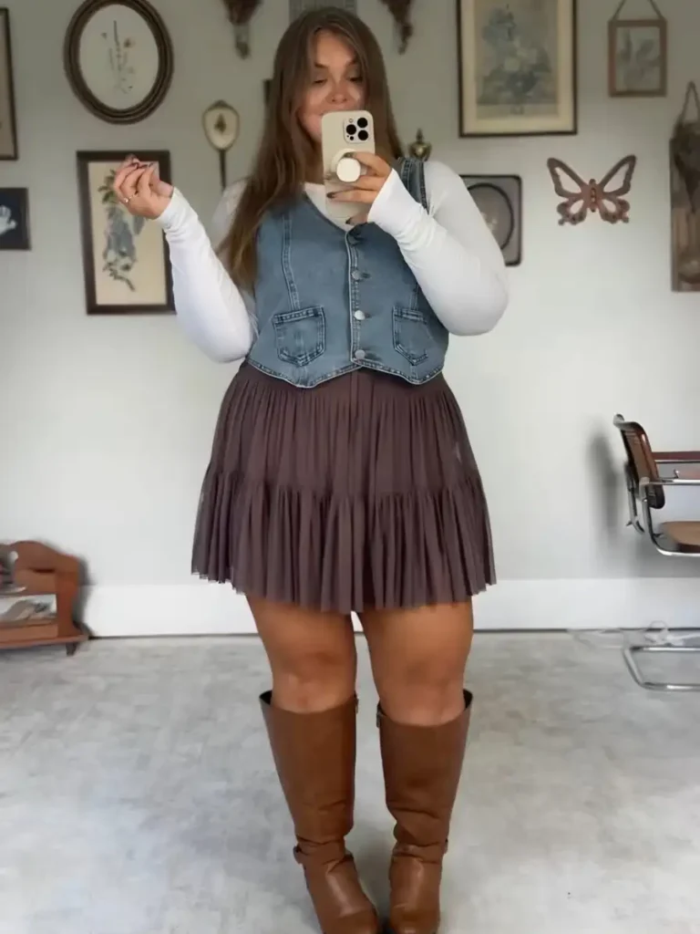 Plus size woman wearing a denim vest over a white long-sleeve top with a brown tiered mini skirt and tall brown boots for a casual fall outfit.