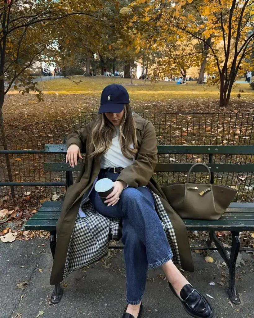 Woman wearing a long olive-brown trench coat with a white top, blue jeans, black loafers, and a navy baseball cap for a relaxed casual fall outfit.