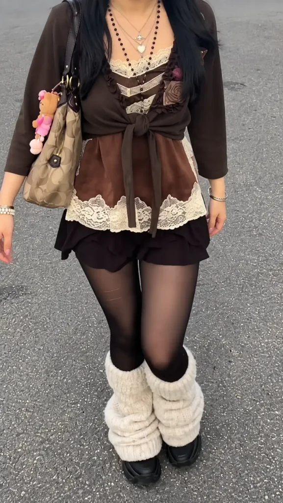 Woman wearing a brown and cream lace corset-style top with a tied brown shrug, dark mini skirt, tights, and cream leg warmers.